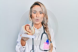Beautiful young blonde doctor woman holding anatomical model of female uterus with fetus scared and amazed with open mouth for