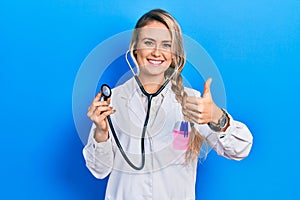 Beautiful young blonde doctor woman holding stethoscope approving doing positive gesture with hand, thumbs up smiling and happy