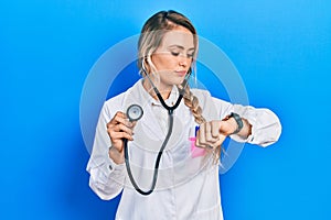 Beautiful young blonde doctor woman holding stethoscope checking the time on wrist watch, relaxed and confident