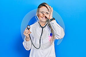 Beautiful young blonde doctor woman holding stethoscope doing ok gesture with hand smiling, eye looking through fingers with happy