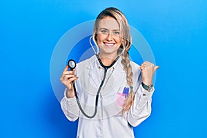 Beautiful young blonde doctor woman holding stethoscope pointing to the back behind with hand and thumbs up, smiling confident