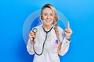 Beautiful young blonde doctor woman holding stethoscope smiling amazed and surprised and pointing up with fingers and raised arms