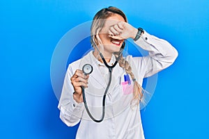 Beautiful young blonde doctor woman holding stethoscope smiling cheerful playing peek a boo with hands showing face