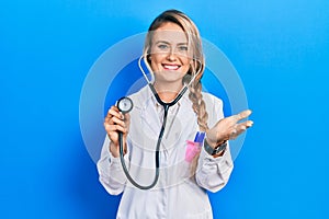 Beautiful young blonde doctor woman holding stethoscope smiling friendly offering handshake as greeting and welcoming