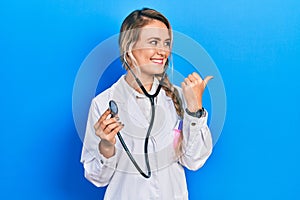 Beautiful young blonde doctor woman holding stethoscope smiling with happy face looking and pointing to the side with thumb up