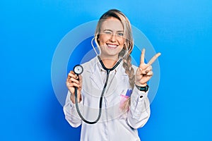 Beautiful young blonde doctor woman holding stethoscope smiling with happy face winking at the camera doing victory sign with