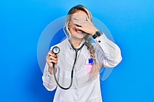 Beautiful young blonde doctor woman holding stethoscope smiling and laughing with hand on face covering eyes for surprise