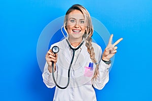 Beautiful young blonde doctor woman holding stethoscope smiling looking to the camera showing fingers doing victory sign