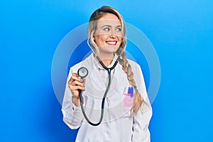 Beautiful young blonde doctor woman holding stethoscope smiling looking to the side and staring away thinking