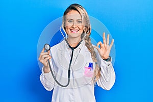 Beautiful young blonde doctor woman holding stethoscope smiling positive doing ok sign with hand and fingers