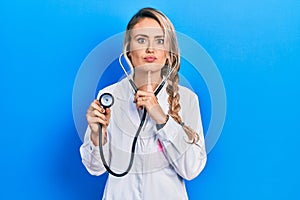 Beautiful young blonde doctor woman holding stethoscope thinking concentrated about doubt with finger on chin and looking up