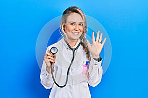 Beautiful young blonde doctor woman holding stethoscope waiving saying hello happy and smiling, friendly welcome gesture