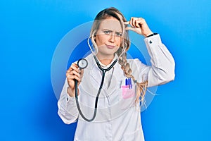 Beautiful young blonde doctor woman holding stethoscope worried and stressed about a problem with hand on forehead, nervous and