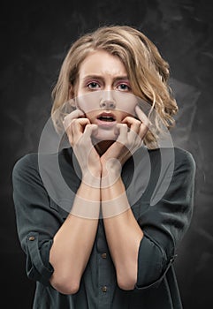 Beautiful young blonde girl with disheveled hairstyle and nude makeup, wearing a shirt and jeans emotionally posing on a gray