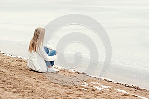 Beautiful young blonde girl in jeans and a white shirt sitting on the shore of the frozen cold of the lake near the forest in earl