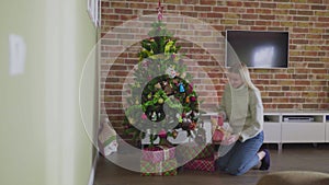 A beautiful young blonde girl puts a gift under the Christmas tree, at home. New Year's Eve. Cozy atmosphere