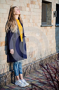 Beautiful young blonde girl in yellow sweater, white sneakers and blue coat, in yellow glasses stay against brick wall