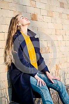 Beautiful young blonde girl in yellow sweater, white sneakers and blue coat, in yellow glasses stay against brick wall