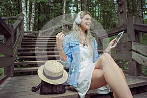 Beautiful young blonde hipster woman listening music in headphones in park with tablet in hands, sitting on wooden steps