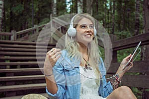 Beautiful young blonde hipster woman listening music in headphones in park with tablet in hands, sitting on wooden steps