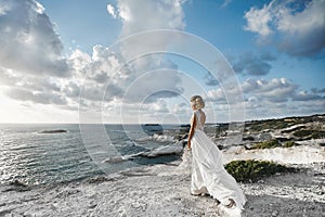 Beautiful young blonde model girl, in white dress, stands half sideways at the coast and looks at the sea