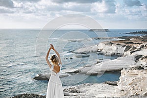 Beautiful young blonde model girl, in white lace dress with naked back, stands with raised hands at the coast and looks