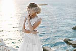 Beautiful young blonde model girl, in white lace dress, stands over the sea coast