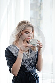 Beautiful young blonde woman in a blue robe by the window. Drinks coffee or tea from a white cup with a saucer. Morning