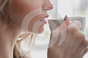 Beautiful young blonde woman in a blue robe by the window. Drinks coffee or tea from a white cup with a saucer. Morning