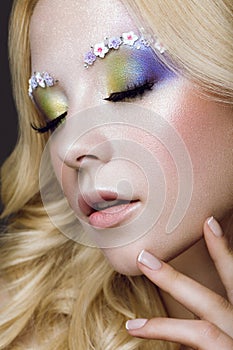 Beautiful young blonde woman with creative make-up color, curls and flowers on eyebrows. Beauty face. Art makeup.