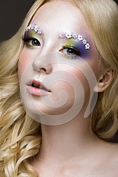 Beautiful young blonde woman with creative make-up color, curls and flowers on eyebrows. Beauty face. Art makeup.