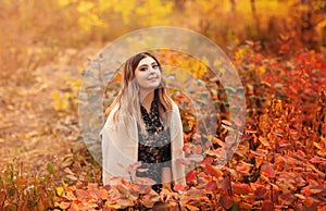 Beautiful young blonde woman in dress sitting with pensive look in tall grass in autumn forest