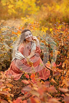 Beautiful young blonde woman in dress sitting with pensive look in tall grass in autumn forest