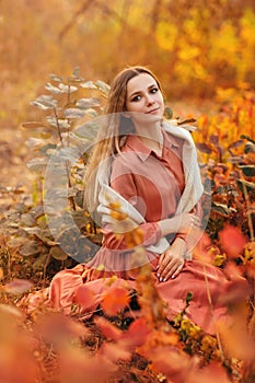 Beautiful young blonde woman in dress sitting with pensive look in tall grass in autumn forest