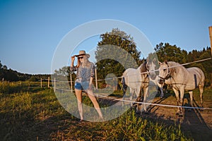 Beautiful young blonde woman dressed safari style in hat and plaid shirt posing with thoroughbreds horses on farm in