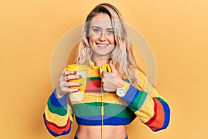 Beautiful young blonde woman drinking cup of coffee wearing headphones doing happy thumbs up gesture with hand