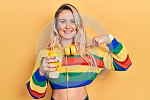 Beautiful young blonde woman drinking cup of coffee wearing headphones looking confident with smile on face, pointing oneself with
