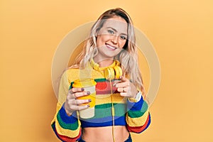 Beautiful young blonde woman drinking cup of coffee wearing headphones pointing fingers to camera with happy and funny face