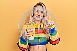 Beautiful young blonde woman drinking cup of coffee wearing headphones pointing to you and the camera with fingers, smiling