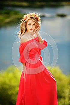 Beautiful young blonde woman with a flower wreath on her head. Beauty girl with flowers hairstyle in a red long dress near the