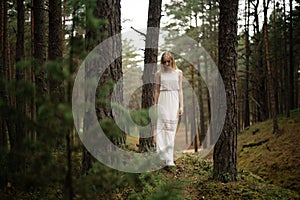 Walking Beautiful young blonde woman forest nymph in white dress in evergreen wood