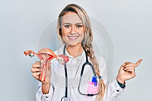 Beautiful young blonde woman holding anatomical model of female genital organ smiling happy pointing with hand and finger to the