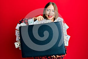 Beautiful young blonde woman holding briefcase full of dollars smiling with a happy and cool smile on face