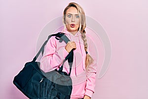 Beautiful young blonde woman holding gym bag scared and amazed with open mouth for surprise, disbelief face