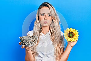 Beautiful young blonde woman holding sunflower seeds an flower puffing cheeks with funny face