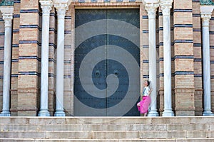 Beautiful young blonde woman leaning on the doorjamb of a large door of a monumental building in the city of Seville. The girl