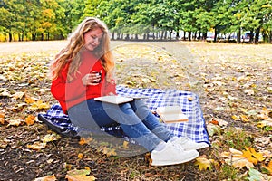 Beautiful young blonde woman with long curly hair reads a book in nature with a cup of tea in her hand. Autumn picnic, outdoor photo