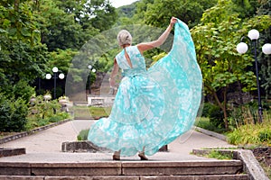 Beautiful young blonde woman in a long green dress is dancing in summer park.