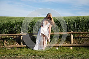 A beautiful young blonde woman with long hair in a wedding dress fastens her high heels while sitting on a wooden fence. In the