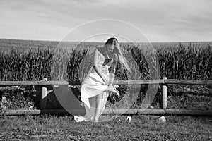 A beautiful young blonde woman with long hair in a wedding dress fastens her high heels while sitting on a wooden fence. In the